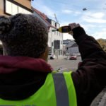 Person in Warnweste misst mit einem Handmessgerät den Verkehr an einer innerstädtischen Straße.