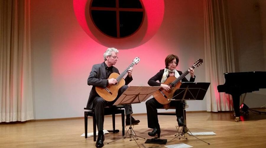 Gitarrenduo auf der Bühne bei einer Veranstaltung 2009 Zwei Gitarristen spielen auf einer Bühne vor einem roten Rundfenster; rechts steht ein schwarzer Konzertflügel.