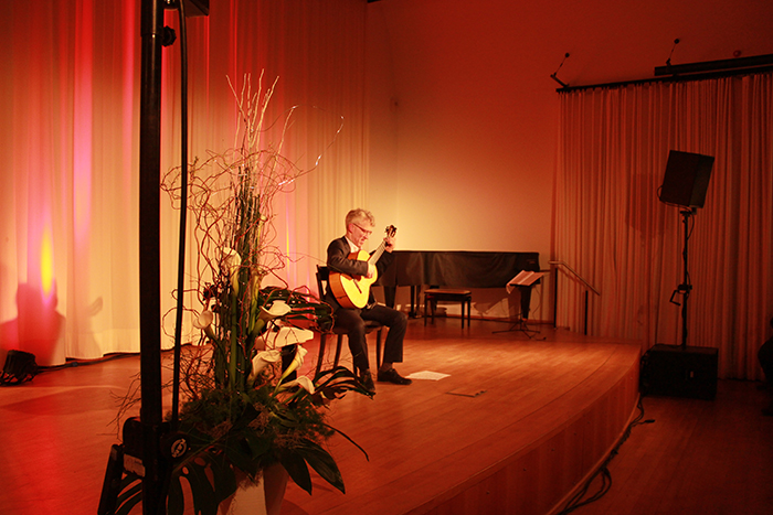 Gitarrist auf der Bühne bei einer Veranstaltung in Deggendorf 2009 Ein sitzender Gitarrist spielt auf einer beleuchteten Bühne neben einem Blumengesteck; im Hintergrund stehen ein Klavier und Lautsprecher.