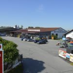 Außenansicht des HolzLand-Hundshammer-Marktgeländes mit Verkaufsgebäuden, Parkplatz und Werbeschildern bei klarem Himmel