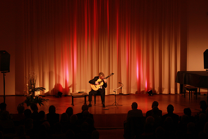 Gitarrist beim Konzert im Jahr 2009 Gitarrist spielt auf einer beleuchteten Bühne vor sitzendem Publikum; rechts steht ein Flügel.