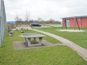 Grasfläche mit Tischtennisplatte, Schaukel und gepflastertem Weg neben einem roten Gebäude; im Hintergrund stehen Bäume und ein Auto.