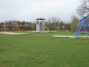 Weiter Spielplatz mit Rasenfläche, Schaukel, Spielturm mit Rutsche und blauem Klettergerät unter bewölktem Himmel