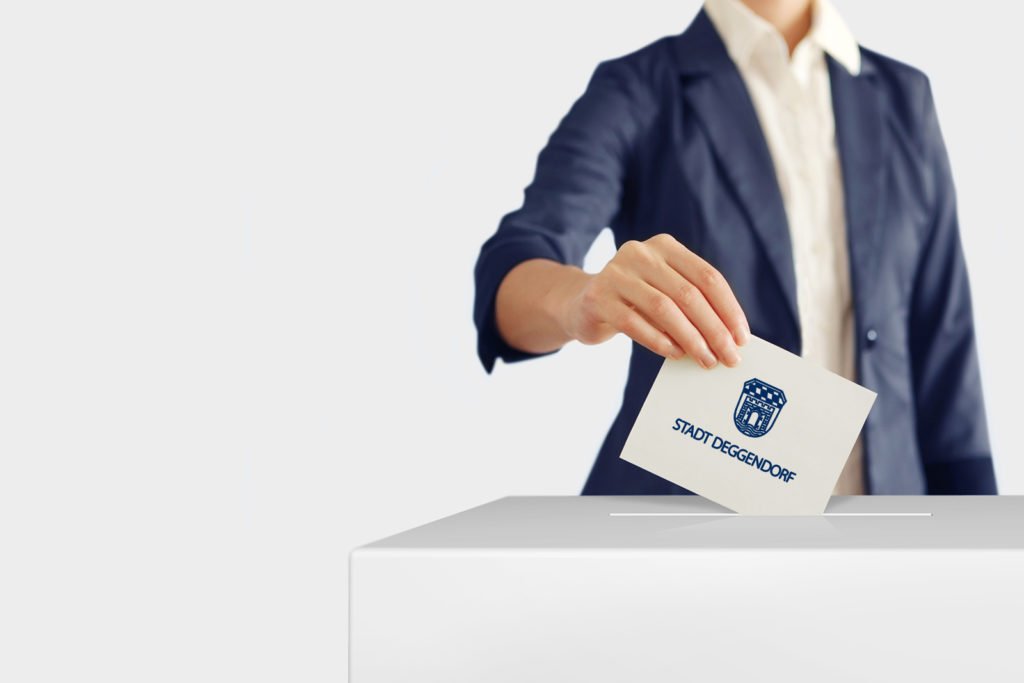 Eine Person steckt einen mit dem Wappen und Schriftzug der Stadt Deggendorf versehenen Wahlumschlag in eine Wahlurne.