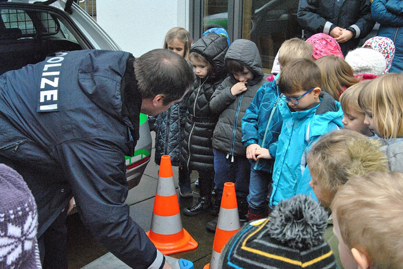 Polizeibeamter zeigt einer Kindergruppe neben einem Polizeifahrzeug Verkehrskegel.
