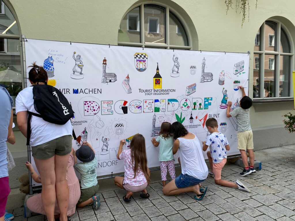 Kinder und Erwachsene gestalten eine große bemalte Wandzeitung mit bunten Motiven und Schriftzügen im Innenhof eines Gebäudes.