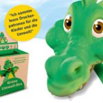 Grüne Drachenfigur neben einer grünen Umwelt-Box für leere Druckerpatronen; ein gelber Hinweis erklärt, dass damit Kinder- und Umweltprojekte unterstützt werden.