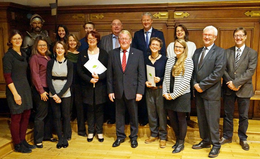 Gruppenfoto von mehreren Erwachsenen bei einer Preisverleihung in einem holzgetäfelten Saal; einige halten Urkunden. Im Hintergrund steht eine historische Rüstung.