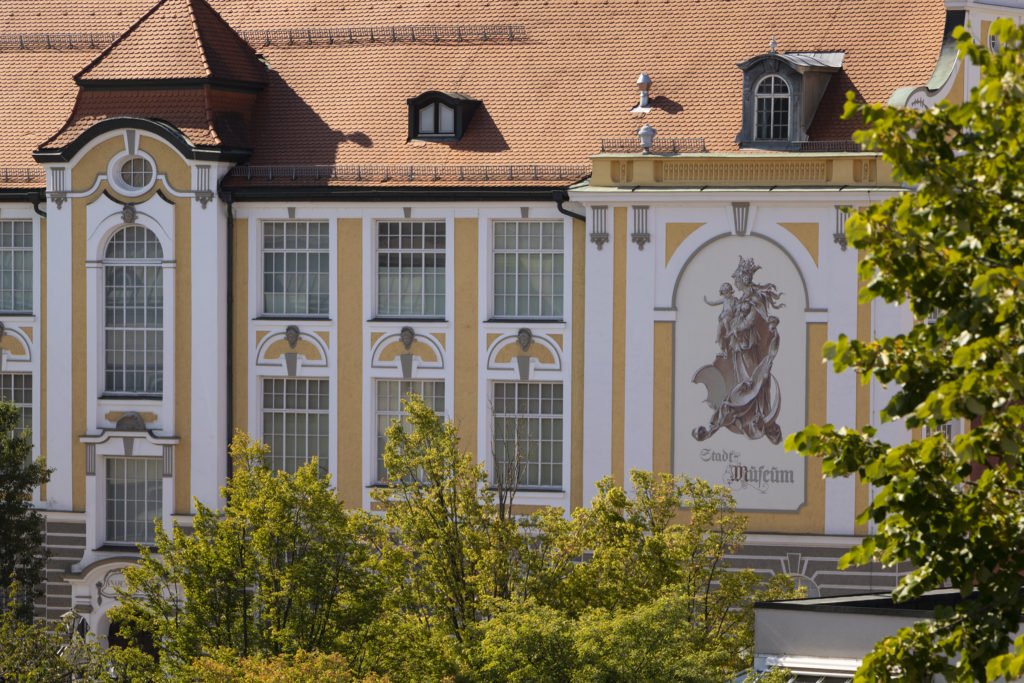 Historische Museumsfassade mit hohen Fenstern, Ornamenten und Wandbild, teilweise von Bäumen verdeckt