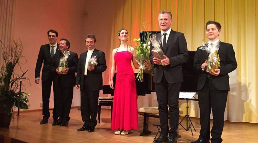 Konzertabend des Vereins in Deggendorf 2009 Sechs festlich gekleidete Personen stehen mit Blumen und Geschenken auf einer Bühne vor einem Klavier und goldfarbenen Vorhängen.