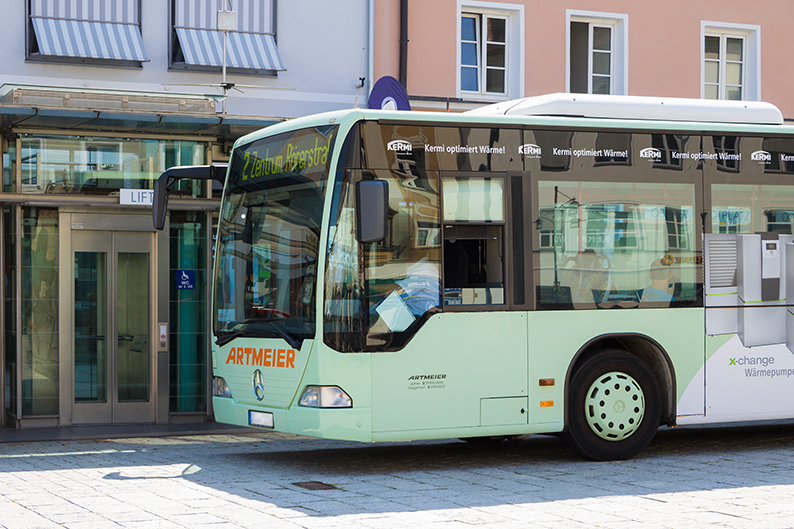 Mintgrüner Stadtbus mit der Aufschrift „ARTMEIER“ steht vor einem Gebäude; im Hintergrund sind Schaufenster und eine barrierefreie Haltestelle zu sehen.
