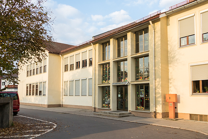 Vorderansicht eines Verwaltungsgebäudes mit Eingang, großen Fenstern und herbstlichen Bäumen