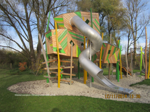 Auffälliger Holzspielplatz mit grünen und orangefarbenen Weiden- und Holzelementen, Kletteraufstieg und großer silberner Röhrenrutsche in einer Parkanlage.