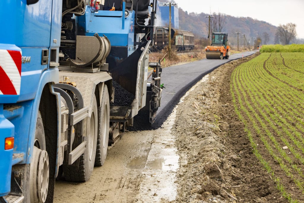Eine Asphaltiermaschine und ein Lastwagen bauen einen Radweg neben einem Feld in Deggendorf.