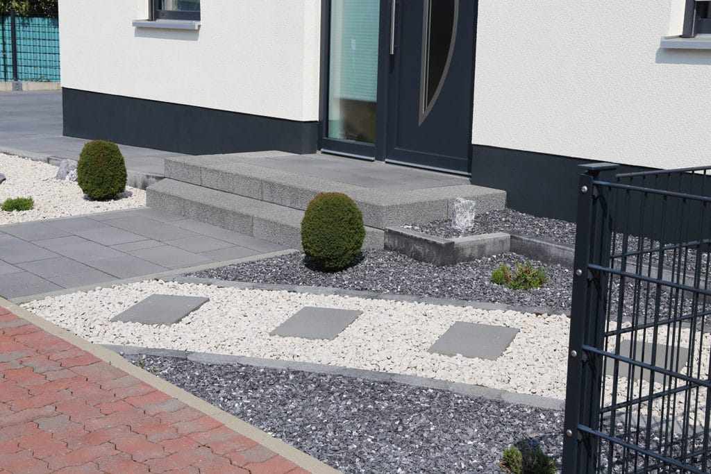 Vorgarten mit weißen und grauen Kiesflächen, Trittplatten, Formschnittsträuchern und Hauseingang