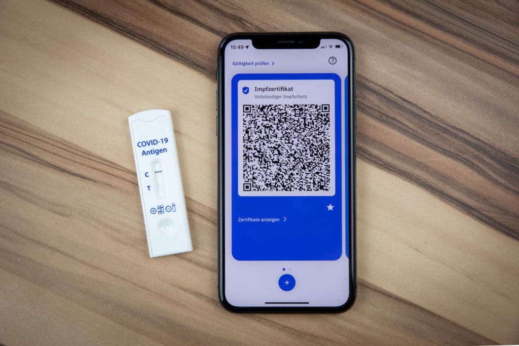 Smartphone mit digitalem Impfzertifikat neben einem COVID-19-Antigentest auf einem Holztisch