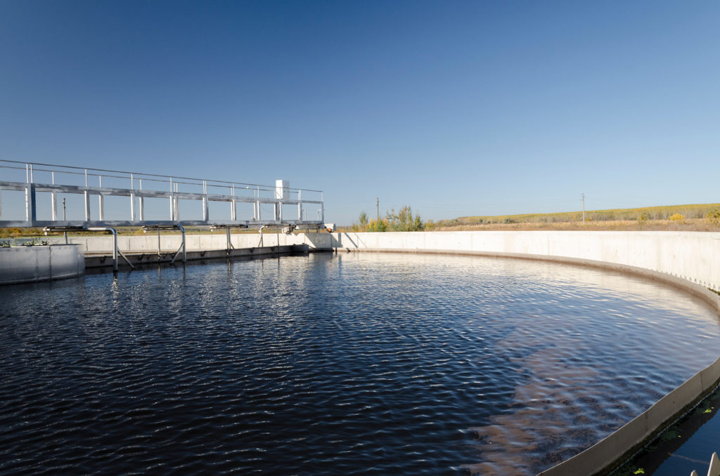 Rundes Absetzbecken einer Kläranlage mit Wasser, Stegen und Metallgeländern vor bewaldeten Hügeln