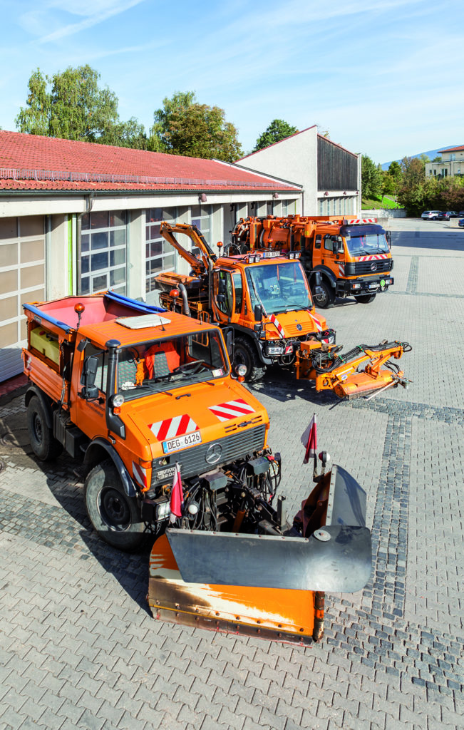 Mehrere orangefarbene Bauhoffahrzeuge mit Schneepflügen und weiteren Anbaugeräten stehen auf dem Gelände des Bauhofs.