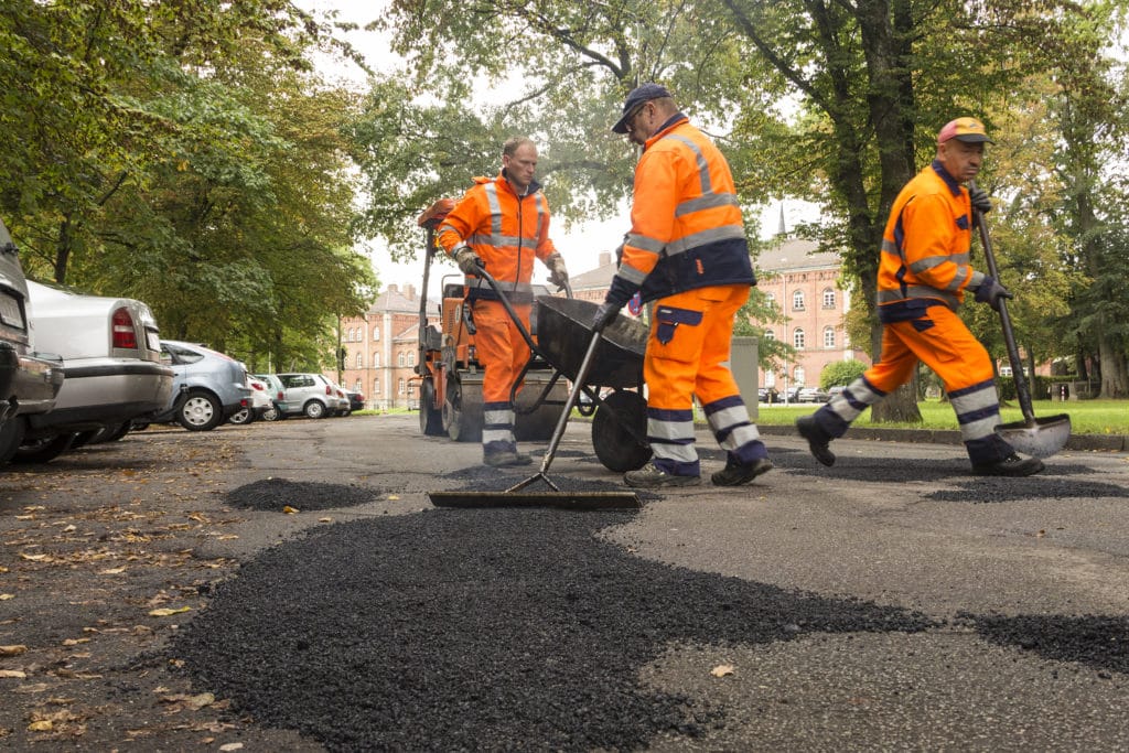 Drei Mitarbeiter in orangefarbener Arbeitskleidung verteilen auf einer Straße frischen Asphalt.