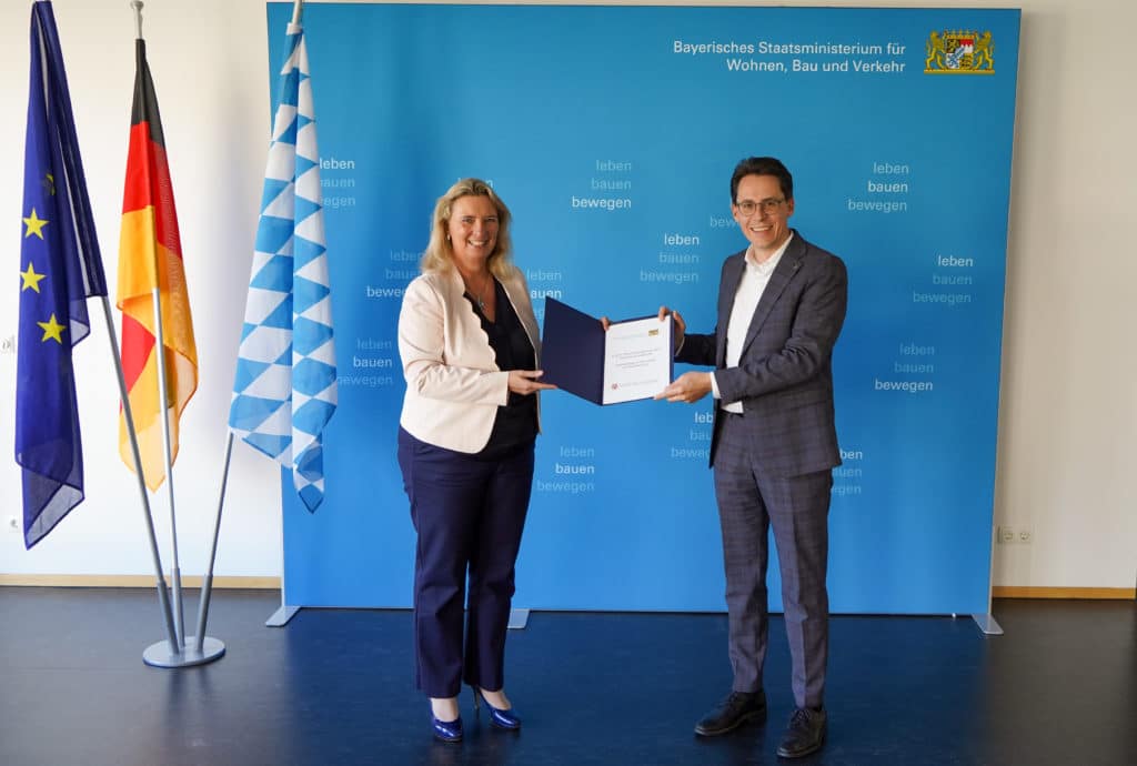 Unterzeichnung einer Vereinbarung zur Sonderbaulast an der Donaubrücke Eine Frau und ein Mann unterzeichnen vor einer blauen Pressewand eine Vereinbarung und halten das Dokument gemeinsam in die Kamera; daneben stehen die Europaflagge, die deutsche Flagge und die bayerische Rautenflagge.