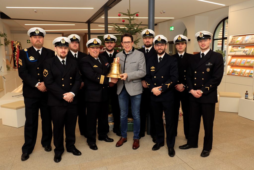 Glockenübergabe Tender Donau 2026 Eine Gruppe von Mitgliedern des Tender Donau steht in Uniform mit einer Person vor einem geschmückten Weihnachtsbaum und hält eine Schiffsglocke.
