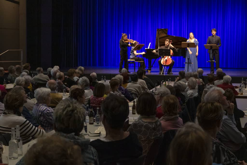 Musikerinnen und Musiker treten vor einem sitzenden Publikum in der blau beleuchteten Stadthalle Deggendorf auf.