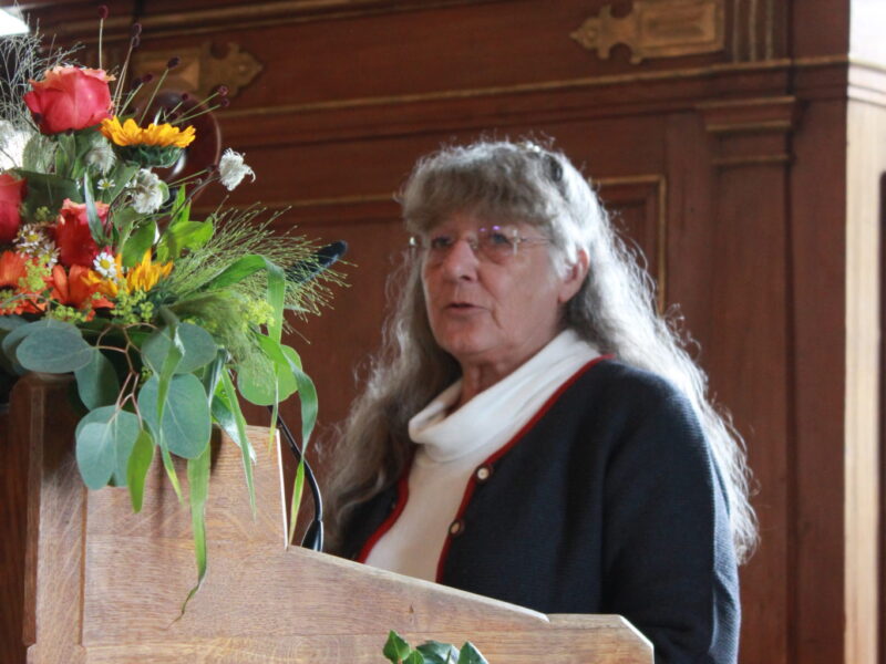 Festrednerin bei einer Veranstaltung in Deggendorf Eine Frau mit langen grauen Haaren spricht an einem holzverkleideten Rednerpult mit Blumenarrangement.