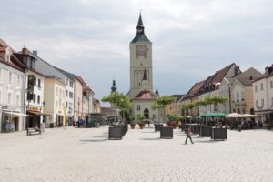 Blick über den Oberen Stadtplatz in Deggendorf auf die Stadtpfarrkirche, flankiert von historischen Häusern, Geschäften und Außengastronomie.