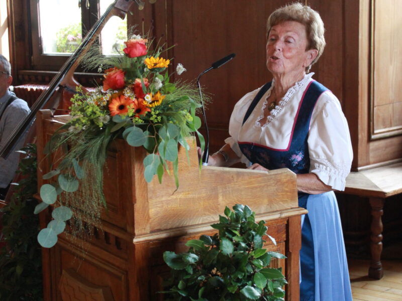 Festrednerin im historischen Rathaus von Deggendorf Eine Frau in bayerischer Tracht spricht an einem hölzernen Rednerpult, das mit Blumen geschmückt ist.