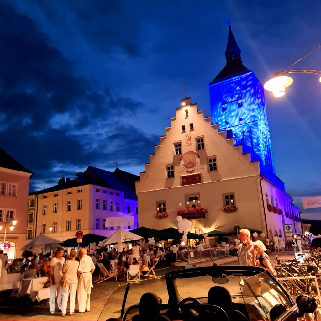 Belebter Marktplatz bei Nacht mit historischem Rathaus, blau beleuchtetem Turm, Gastronomieständen und zahlreichen Besucherinnen und Besuchern