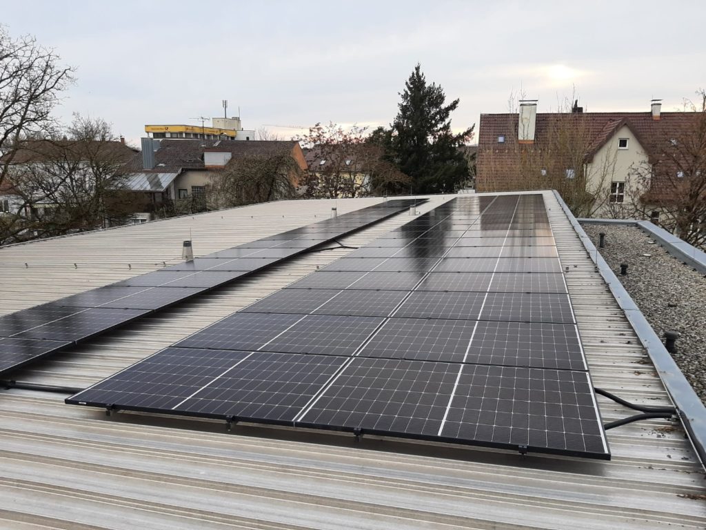 Reihen von Solarmodulen auf einem flachen Schuldach in Deggendorf; im Hintergrund sind Wohnhäuser und Bäume zu sehen.