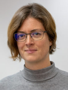 Porträt einer Frau mit schulterlangem braunem Haar, Brille und grauem Rollkragenpullover vor hellem Hintergrund.