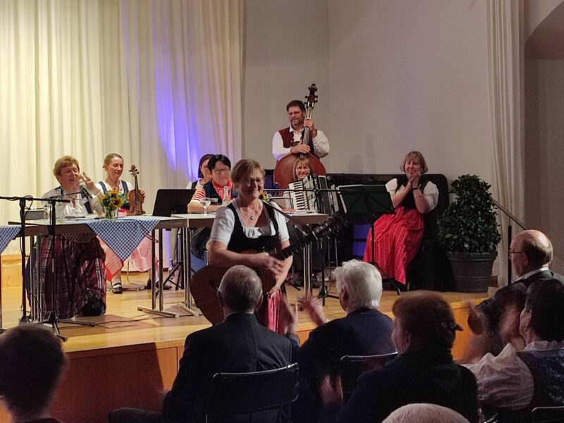 Musikveranstaltung des Vereins M23 in Deggendorf Musikerinnen und Musiker treten vor Publikum in einem festlich dekorierten Saal auf; eine Sängerin steht im Vordergrund, begleitet von weiteren Instrumentalisten.