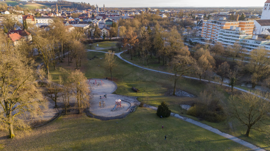 Stadtpark im Frühjahr 2023 Luftaufnahme eines Stadtparks mit verschlungenen Wegen, Spielplatz, großen Bäumen und Wohnhäusern im Hintergrund.