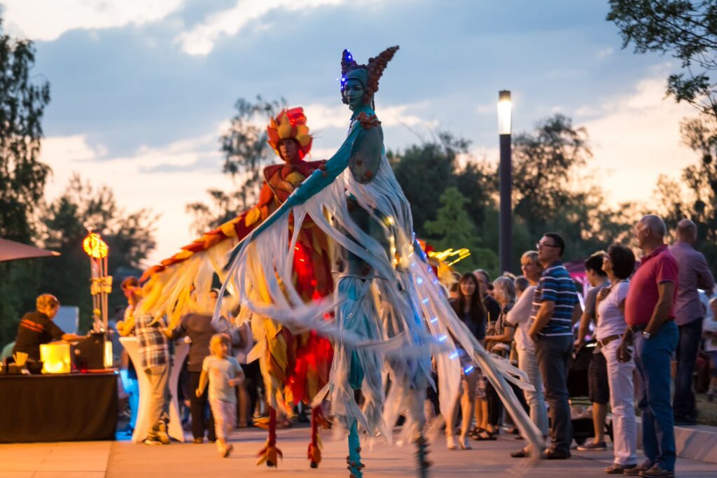 Auftritt beim Donaufest in Deggendorf Zwei farbenfroh kostümierte Stelzenläufer mit leuchtenden Dekorationen treten vor einem Publikum im Freien auf.