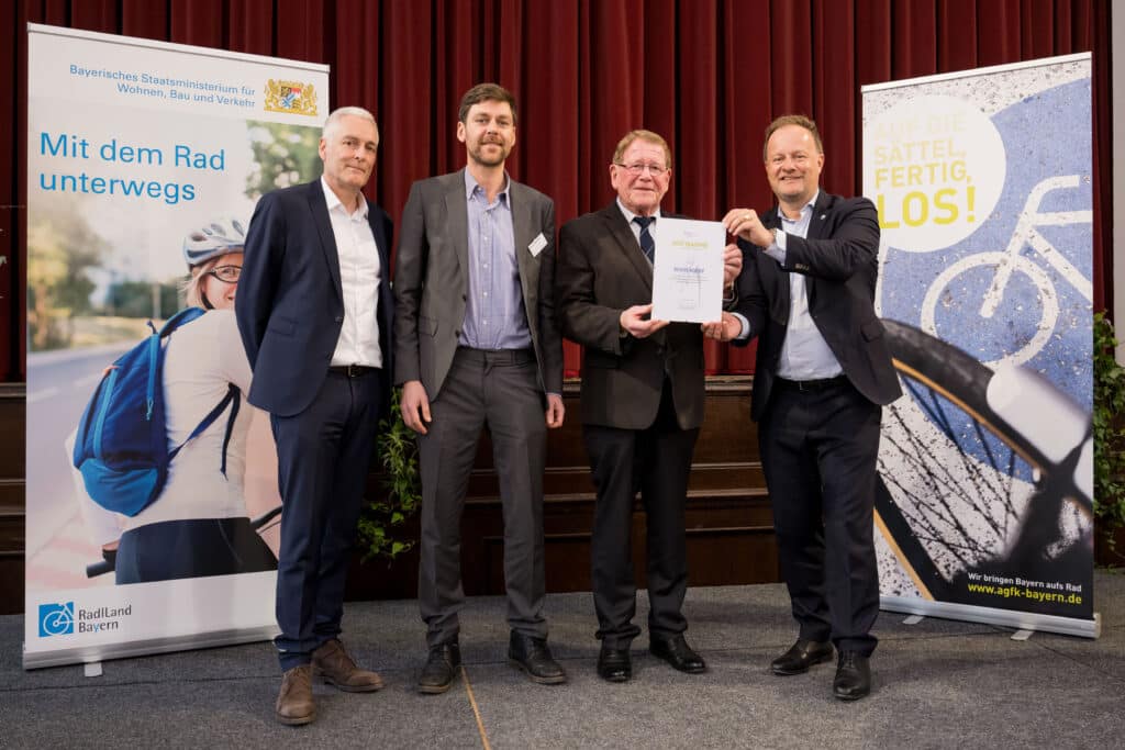 Deggendorf wird Mitglied der AGFK Bayern Vier Männer stehen auf einer Bühne vor Fahrradmotiven; zwei von ihnen halten eine Urkunde zur Mitgliedschaft in der AGFK Bayern.