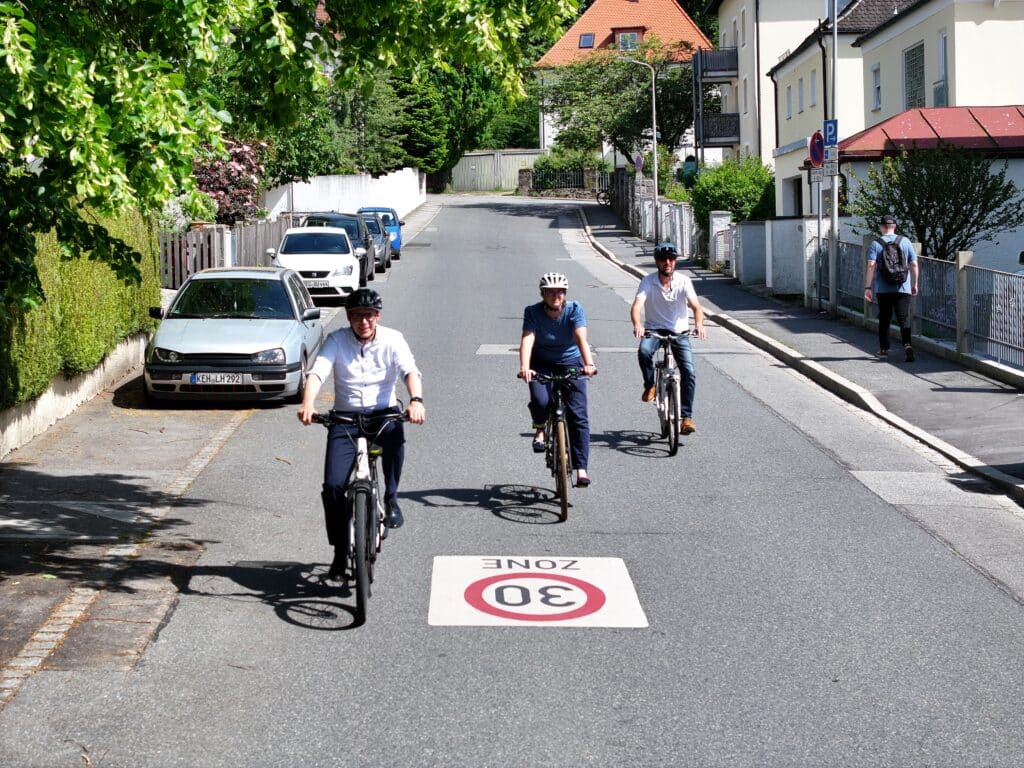 Drei Radfahrende fahren durch eine von Häusern gesäumte Straße; auf der Fahrbahn ist eine 30-Zone markiert.