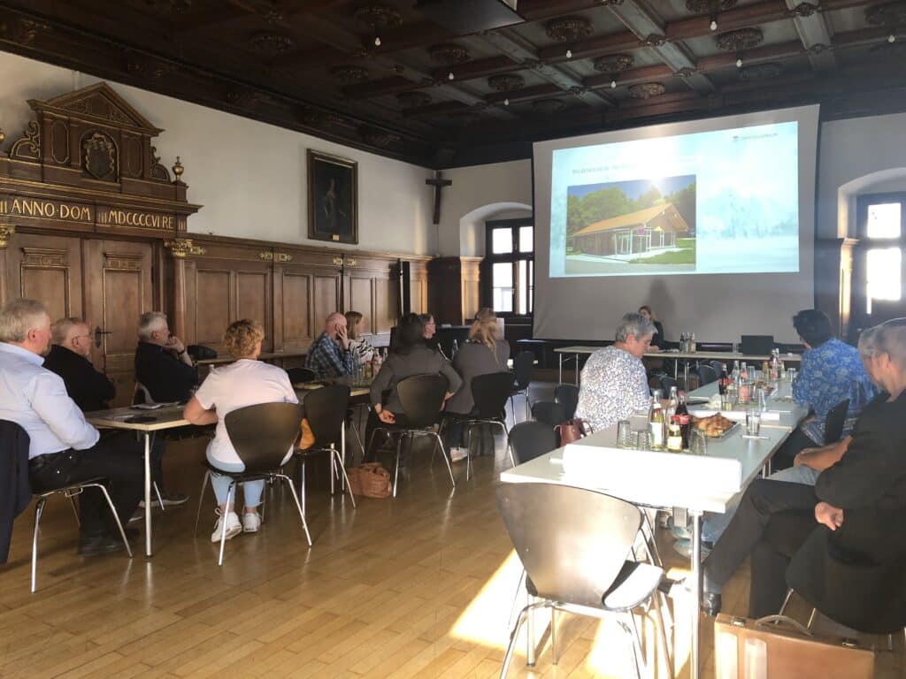 Teilnehmende sitzen bei einer Präsentation in einem historischen Saal; auf der Leinwand sind Landschafts- und Gebäudeaufnahmen zu sehen.
