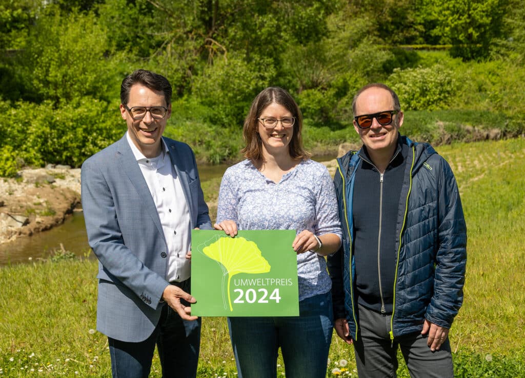 Umweltpreis 2024: Auszeichnung am Gewässer Drei Personen stehen an einem bewachsenen Gewässer und halten gemeinsam ein grünes Schild mit der Aufschrift „Umweltpreis 2024“ in die Kamera.