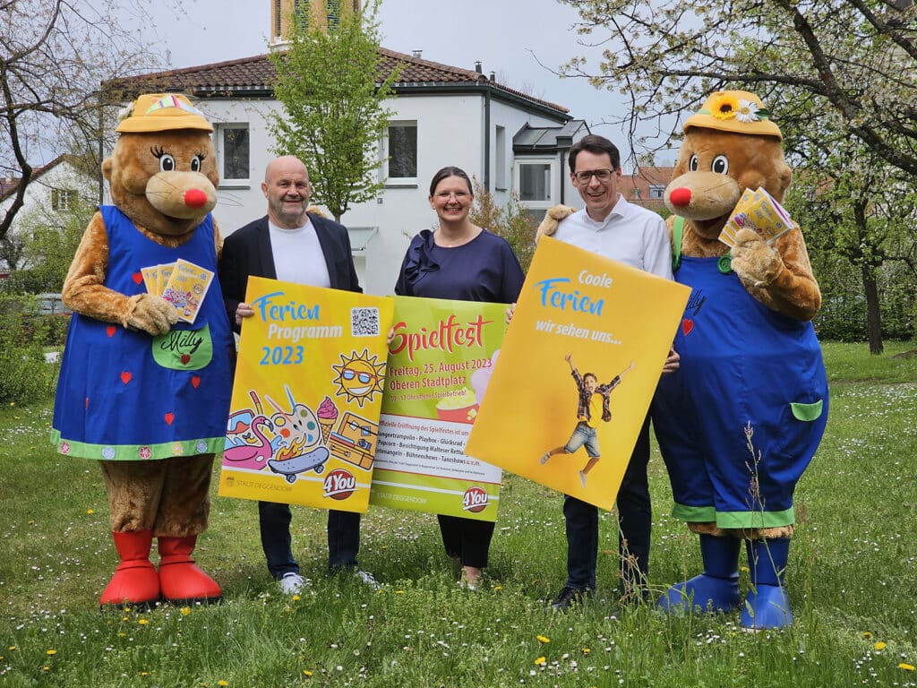 Zwei als Bären verkleidete Figuren und drei Erwachsene präsentieren im Garten bunte Plakate zum Ferienprogramm und Jugendfest.