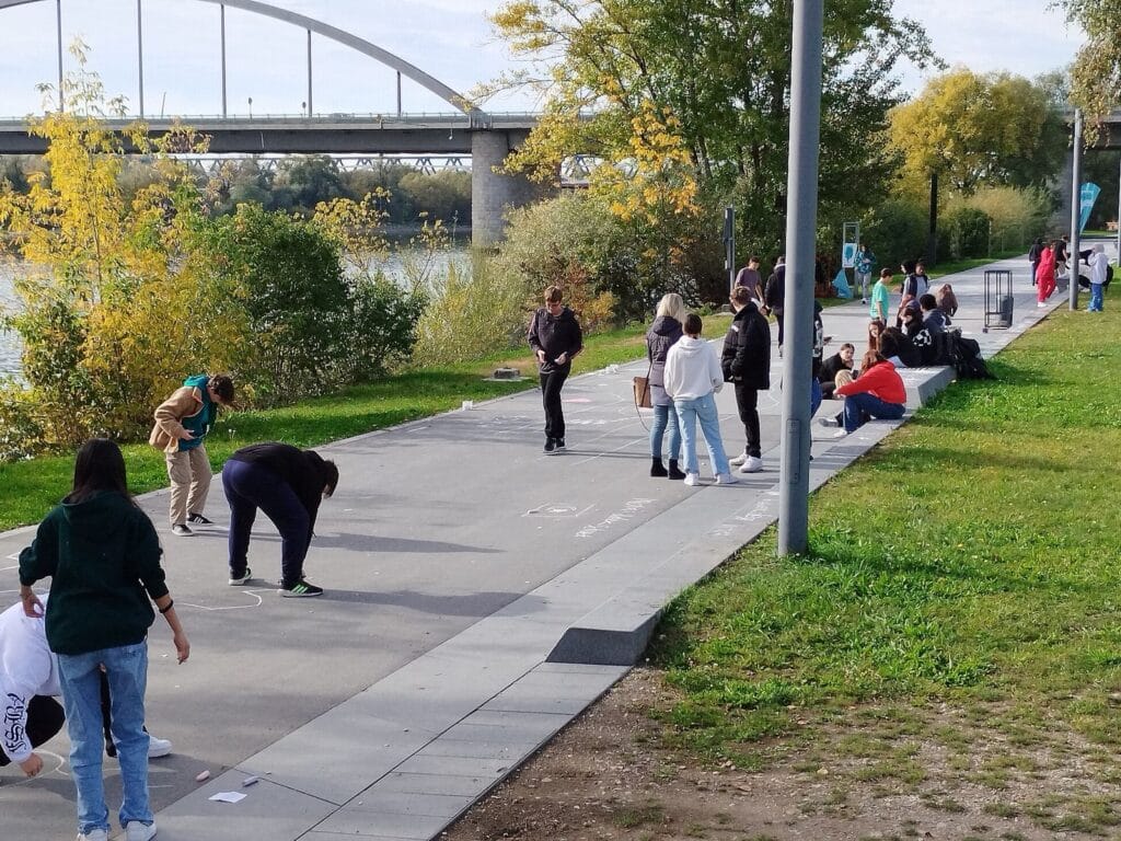 Jugendliche und junge Erwachsene treffen sich auf einem Weg am Rheinufer; im Hintergrund sind eine Brücke, Bäume und Sitzgelegenheiten zu sehen.