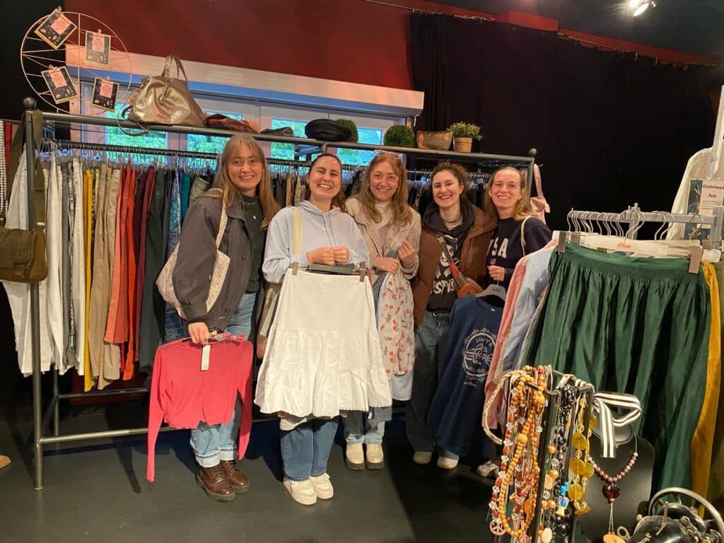 Fünf Frauen stehen bei einer Kleidertauschparty vor Kleiderständern mit gebrauchter Kleidung und Accessoires.