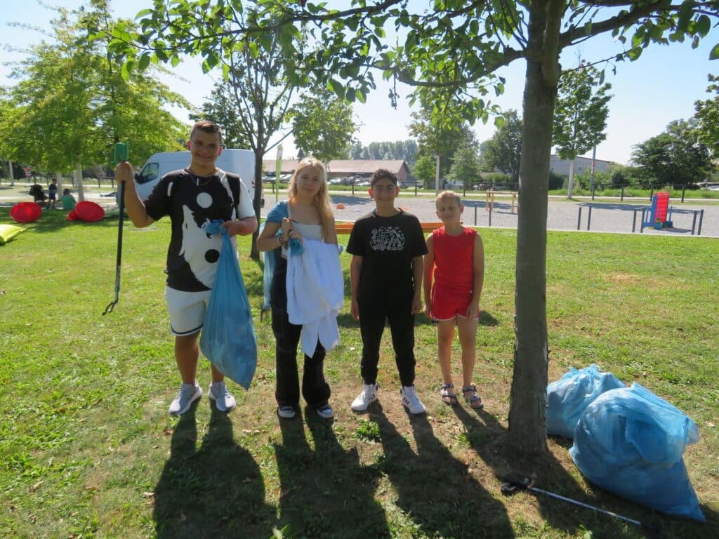 4You beim Weltaufräumtag im September 2024 Vier junge Menschen stehen mit einem blauen Müllsack unter einem Baum auf einer Wiese; weitere gefüllte Müllsäcke liegen daneben.