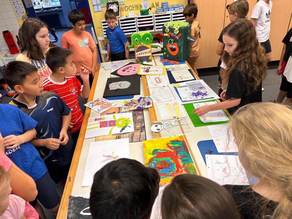 Kinder betrachten eine Ausstellung mit selbst gestalteten Bildern, Bastelarbeiten und Lernplakaten auf mehreren Tischen in einem Klassenraum.