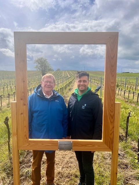 Stadtratsfahrt nach Neusiedl am See 2024 Zwei Männer stehen in einem großen Holzrahmen vor einem Weinberg unter bewölktem Himmel.