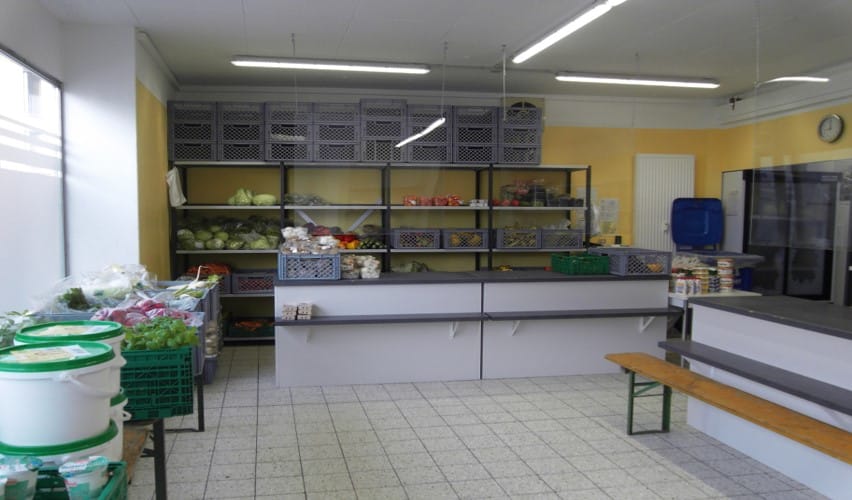 Innenraum einer Tafel-Ausgabestelle mit Regalen voller Obst, Gemüse und weiteren Lebensmitteln sowie Ausgabetresen und Sitzbank.