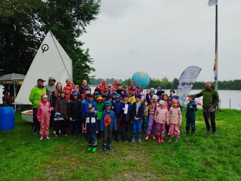 Piratentag 2025 am See Kinder und Erwachsene stehen bei regnerischem Wetter auf einer Wiese am See neben einem weißen Segel und einem bunten aufblasbaren Ball.