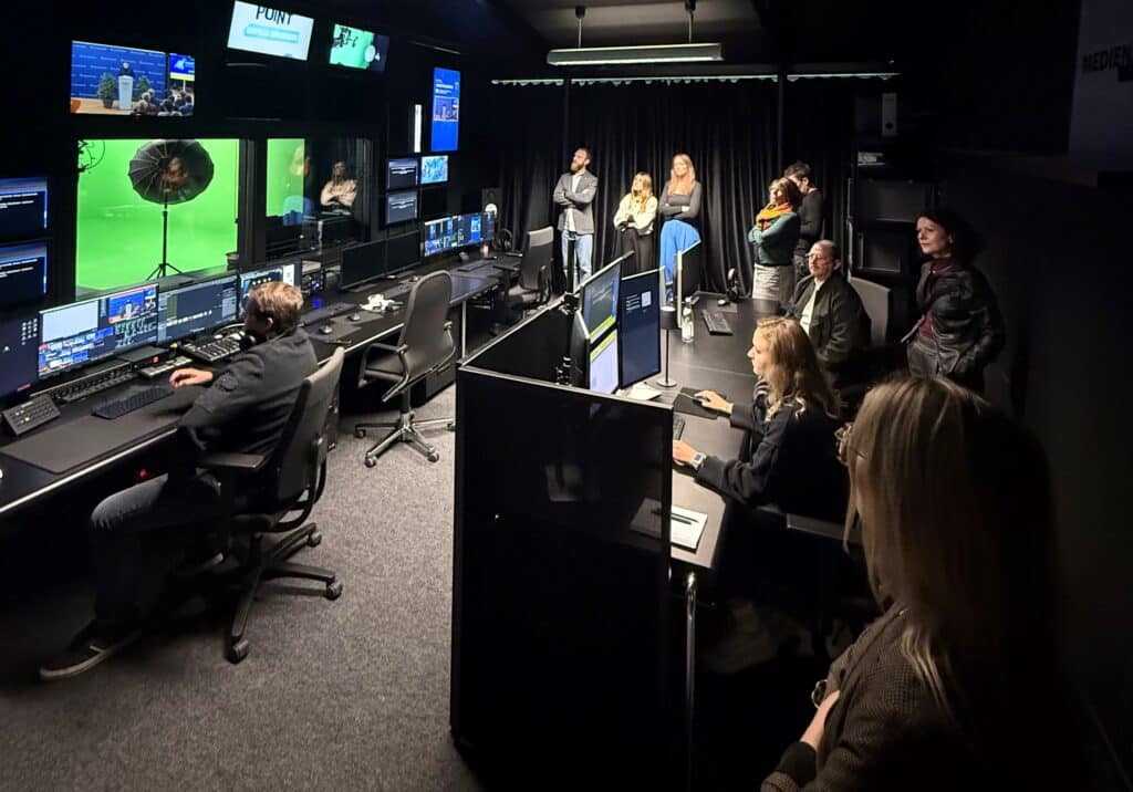 Menschen arbeiten in einem Medienregieraum mit mehreren Monitoren, Kameratechnik und einer grünen Studiofläche; im Hintergrund stehen weitere Personen.