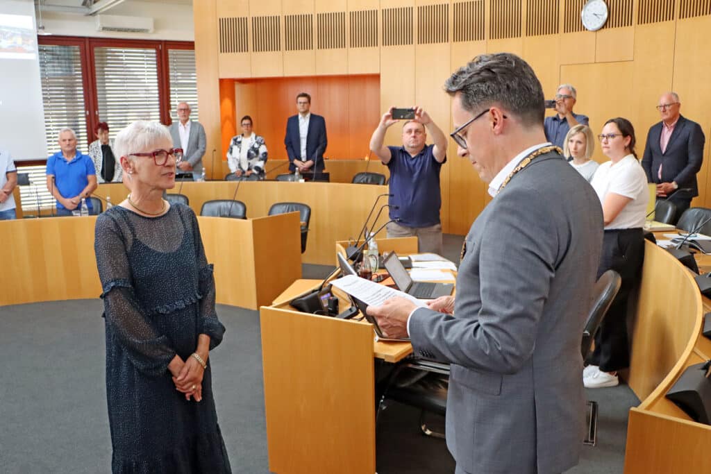 Ein Mann im grauen Anzug spricht am Rednerpult mit einer Frau in einem dunklen Kleid; Mitglieder und Gäste sitzen und stehen im Sitzungssaal.