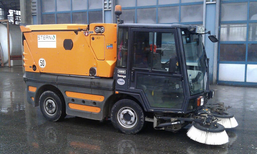Orangefarbene Kehrmaschine mit zwei Rundbürsten steht auf einem nassen Betriebshof.